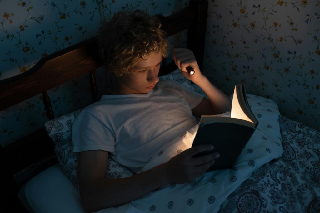 A teenager reads a book under the blanket with a flashlight in a cozy bedroom.