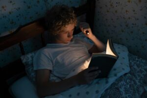 A teenager reads a book under the blanket with a flashlight in a cozy bedroom.